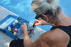 A woman with blonde hair tests pool water using a chemical kit, surrounded by colorful bottles and tools near a clear blue swimming pool.